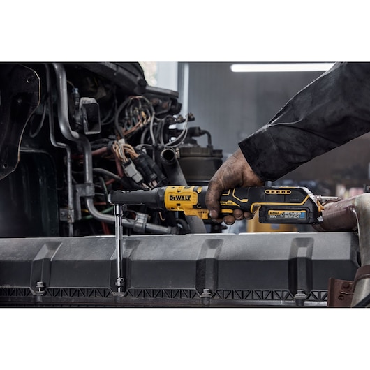 A person uses a DEWALT cordless ratchet tool, model DCF510N, to work on an engine in a workshop. The tool is yellow and black, and the user's hand is dirty from mechanical work.
