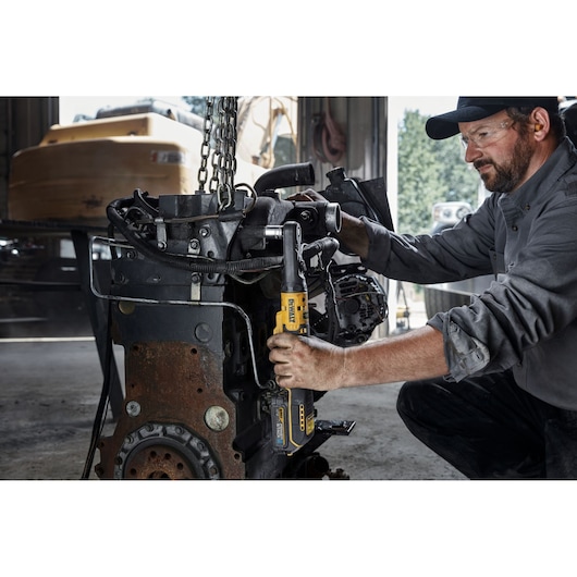 A person using a DEWALT DCF510N cordless ratchet tool to work on a large metal engine component in a workshop. The person's face is blurred for privacy, and the workshop setting includes various tools and machinery.