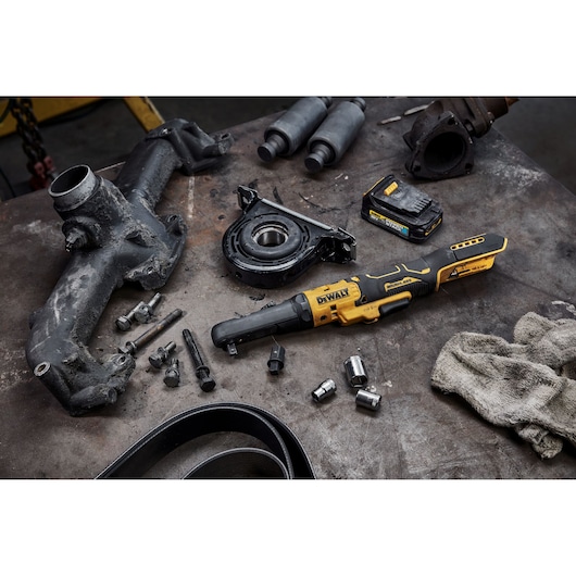 A DEWALT cordless ratchet tool model DCF510N is shown on a workbench alongside various automotive parts, bolts, sockets, a battery pack, and a dirty cloth. The surface of the workbench is scattered with mechanical components, suggesting an ongoing repair or maintenance task.