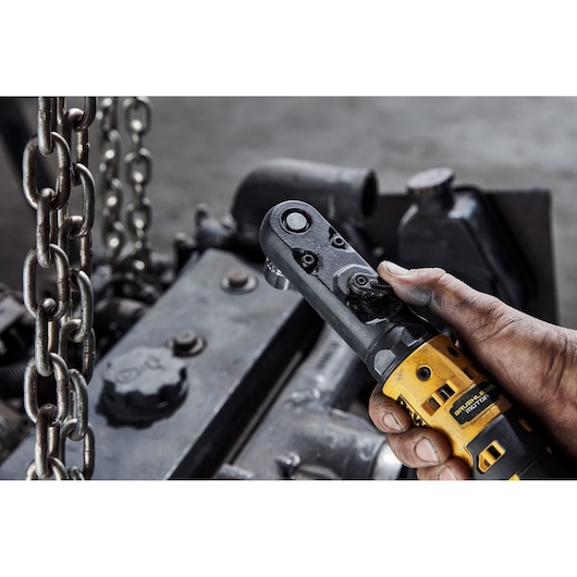 A close-up of a hand holding a DEWALT cordless ratchet tool, model DCF510N, being used in an industrial setting near metal chains and machinery parts.