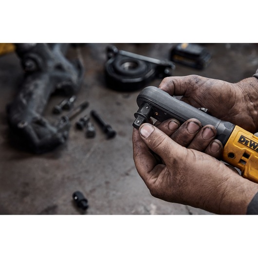 Close-up of a person with dirty hands holding a DEWALT cordless ratchet tool, surrounded by bolts and mechanical parts on a workbench.