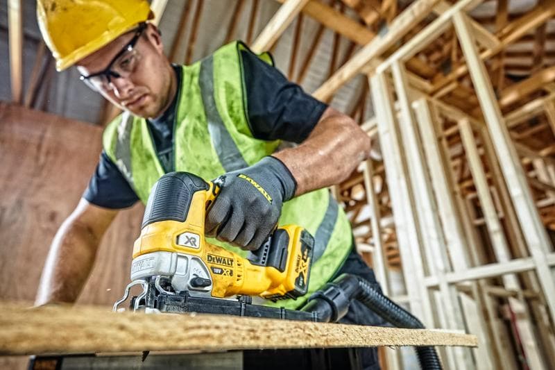 A person wearing a yellow hard hat, safety vest, and gloves is using a DEWALT DCS334P2 cordless jigsaw to cut a wooden board at a construction site with unfinished wooden framing.