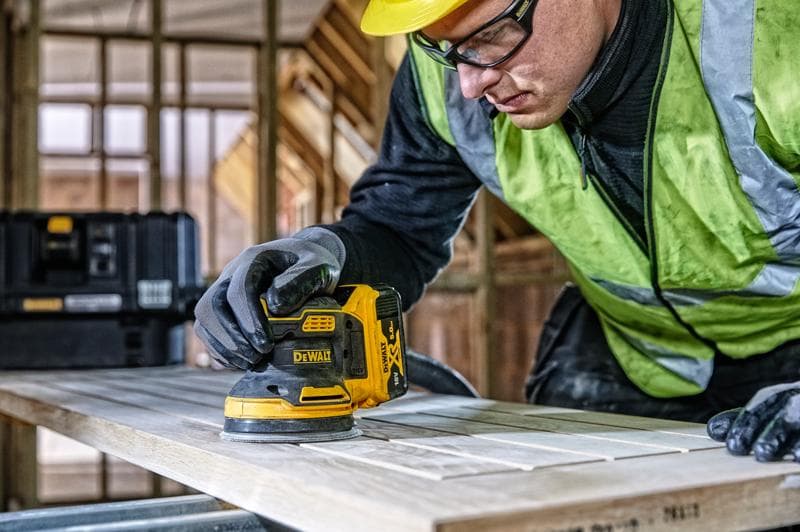 A person wearing work gloves and a high-visibility vest uses a DEWALT cordless palm sander (model DCW210) to sand a wooden board in a construction site. A black DEWALT toolbox is visible in the background.