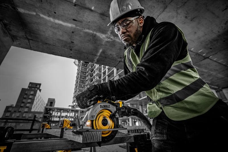 Person using DEWALT DCS565NT cordless circular saw at a construction site, wearing safety gear and high-visibility vest.