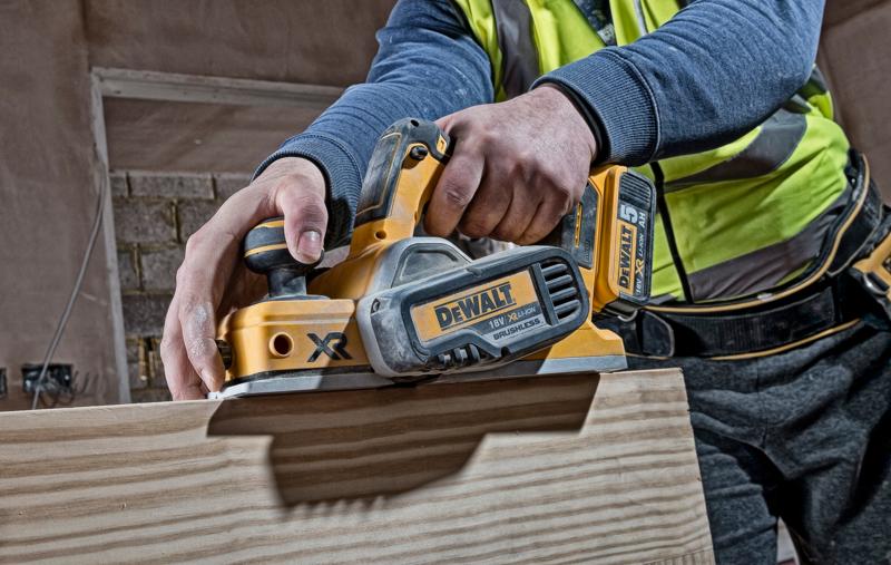 A person using a DEWALT DCP580N cordless planer to smooth the surface of a wooden board. The tool is yellow and black, with visible branding and battery.