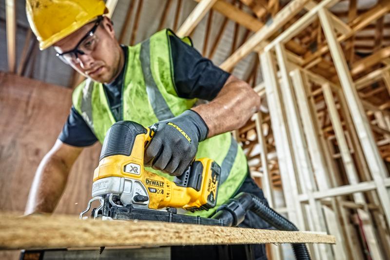 A person wearing a yellow hard hat, safety vest, and gloves is using a DEWALT DCS334P2 cordless jigsaw to cut a wooden board at a construction site with unfinished wooden framing.