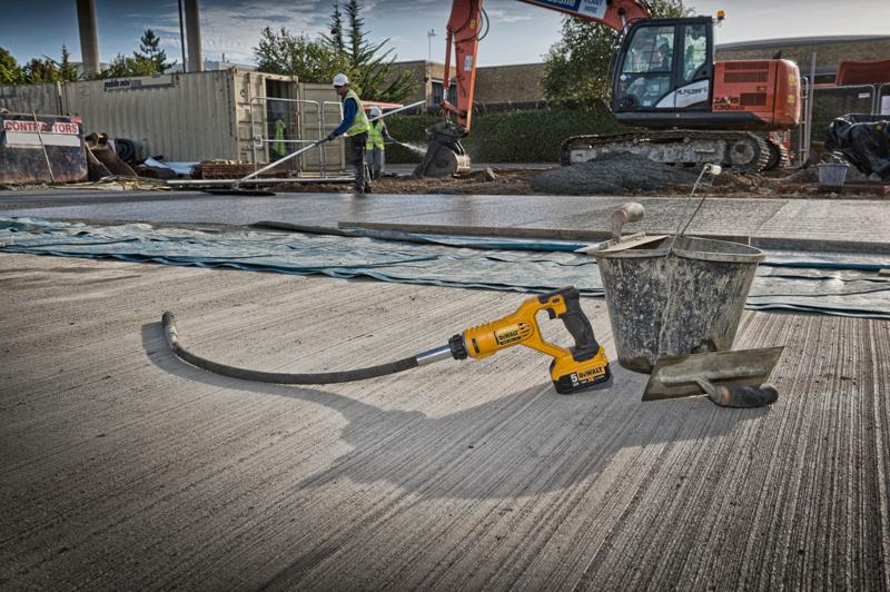 A DEWALT DCE531N concrete vibrator tool is placed on a construction site, next to a metal bucket and a trowel on a freshly laid concrete surface. In the background, there are construction workers, a large excavator, and other building materials.