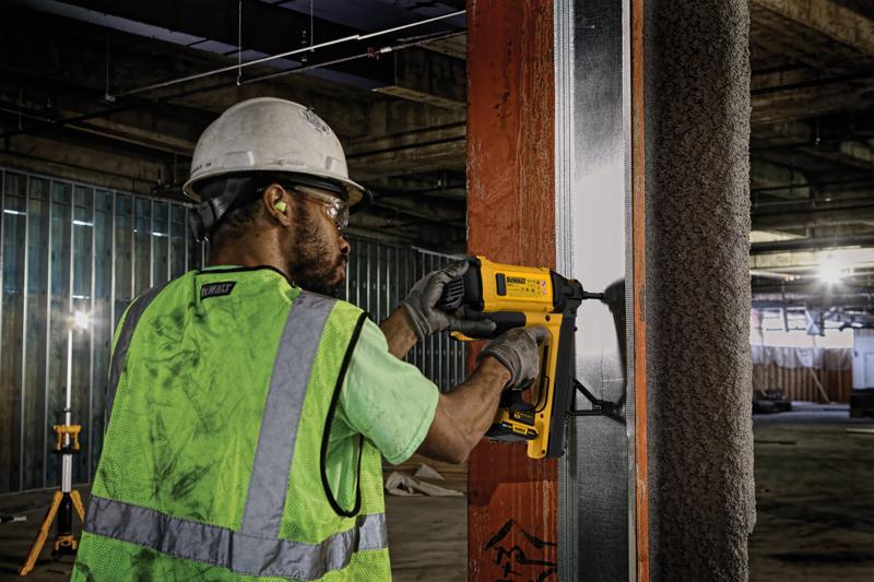 A construction worker wearing a safety helmet, ear protection, gloves, and a high-visibility vest uses a DEWALT nail gun to secure metal framing to a steel beam at an indoor construction site.