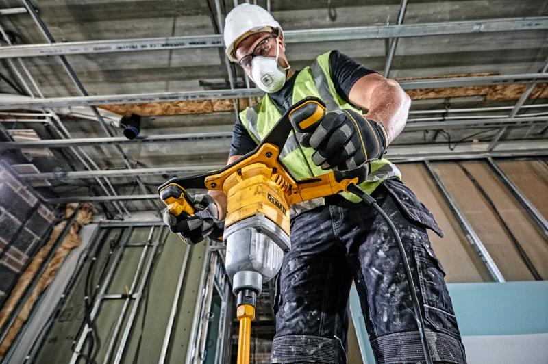 A construction worker wearing a safety vest, gloves, and a hard hat is operating a DEWALT DWD241 electric drill mixer on a building site, surrounded by metal framework and unfinished walls.